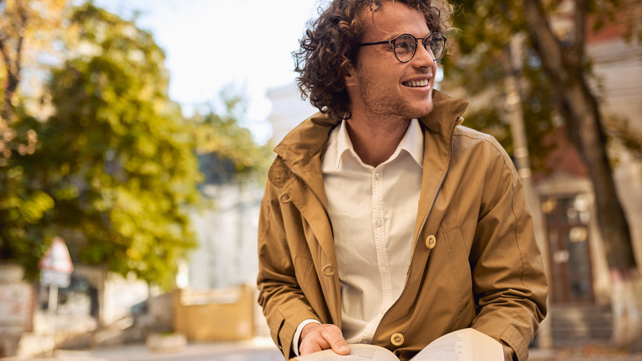 Glücklicher junger Mann mit Brille und lockigem Haar liest und posiert mit Buch im Freien